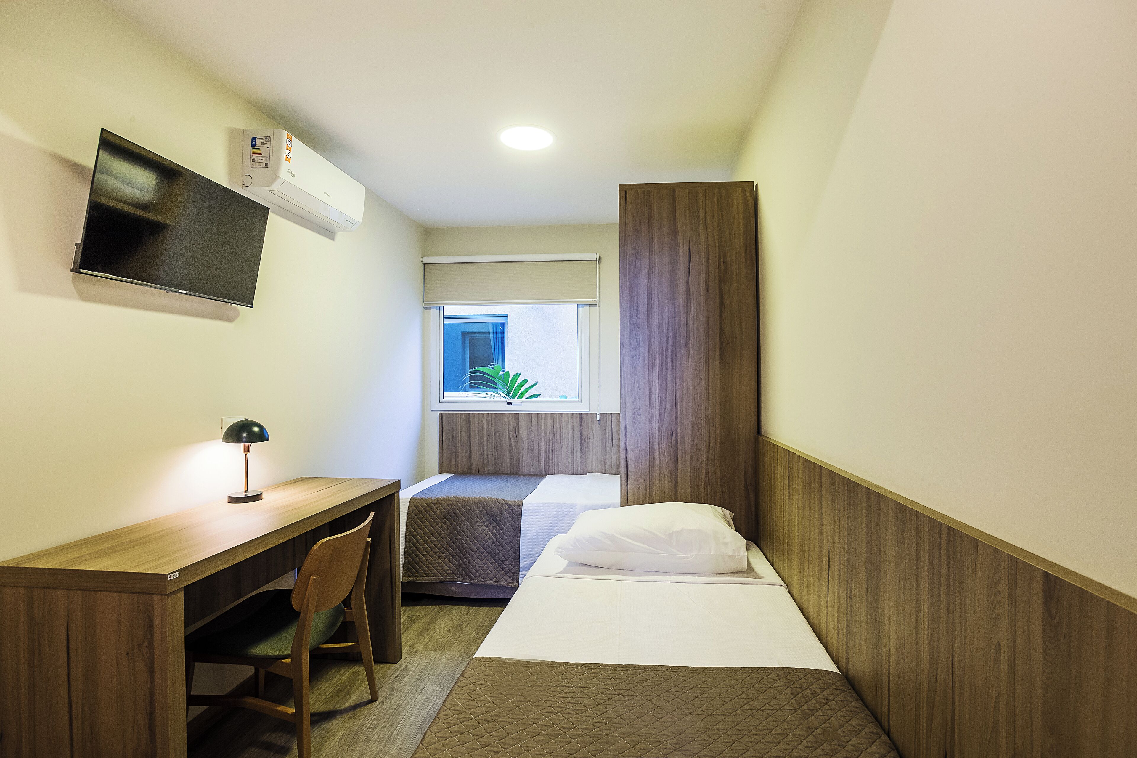 Habitación de hotel compacta con escritorio de madera, una cama individual, mesita de noche pequeña, televisión montada en la pared y ventana con vistas a zonas verdes.