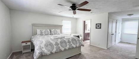 Un dormitorio con cama, mesita de noche, ventilador de techo y una puerta que da a otra habitación.