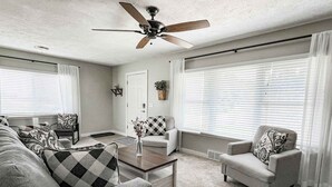 Un salon avec un ventilateur de plafond, un canapé, un fauteuil, une table basse et une fenêtre avec des stores.