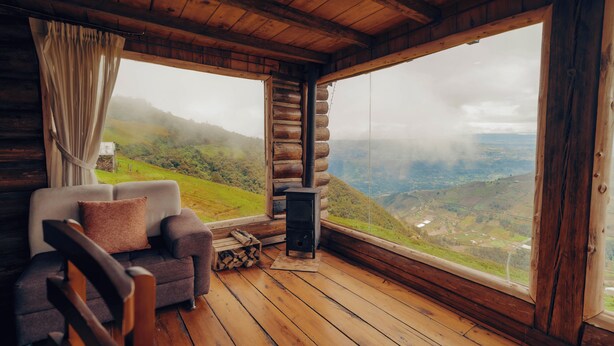 Interior de una cabaña de troncos con sofá, chimenea y un ventanal con vistas a un valle.