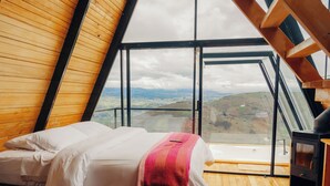 Una habitación con techos altos y una inclinada cubierta de madera, una cama con ropa blanca y una manta rosa, piso de madera, y un ventanal con vistas al campo.