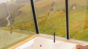 Un baño moderno con una bañera grande, un taburete de madera y vistas a un paisaje verde a través de amplios ventanales.