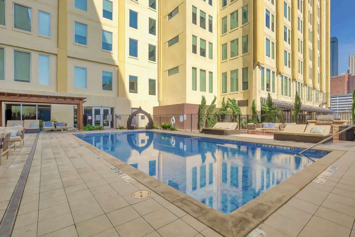 A hotel pool area with a clear view of the pool, lounge chairs, and a building with multiple floors and windows.