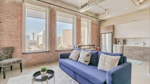 Un salon moderne avec un canapé bleu, une petite table ronde agrémentée d’une plante, et une vue sur la ville grâce à de grandes fenêtres.