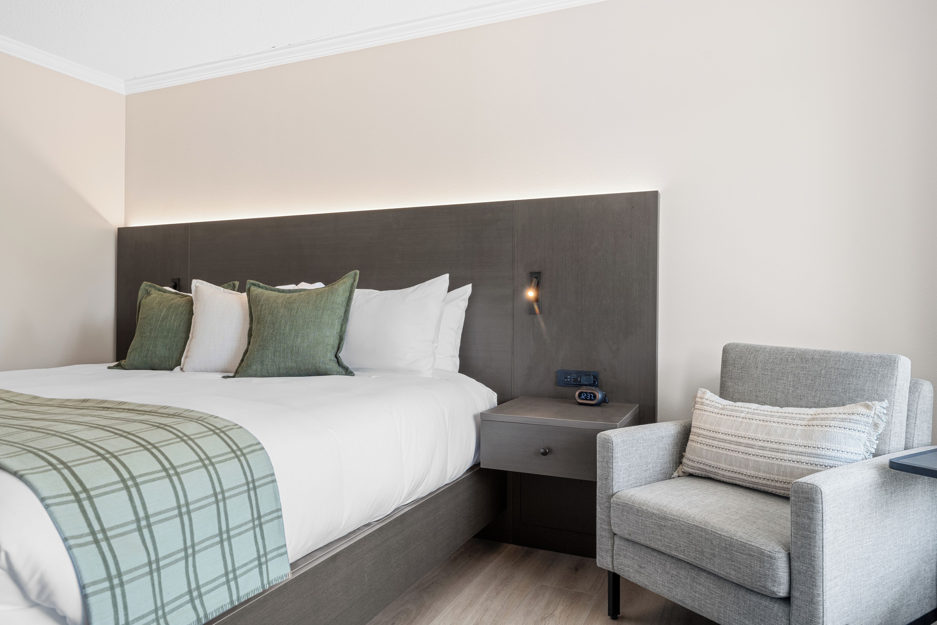 Een moderne hotelkamer met een groot bed, een nachtkastje met een klok, een grijze fauteuil en een wandlamp.
