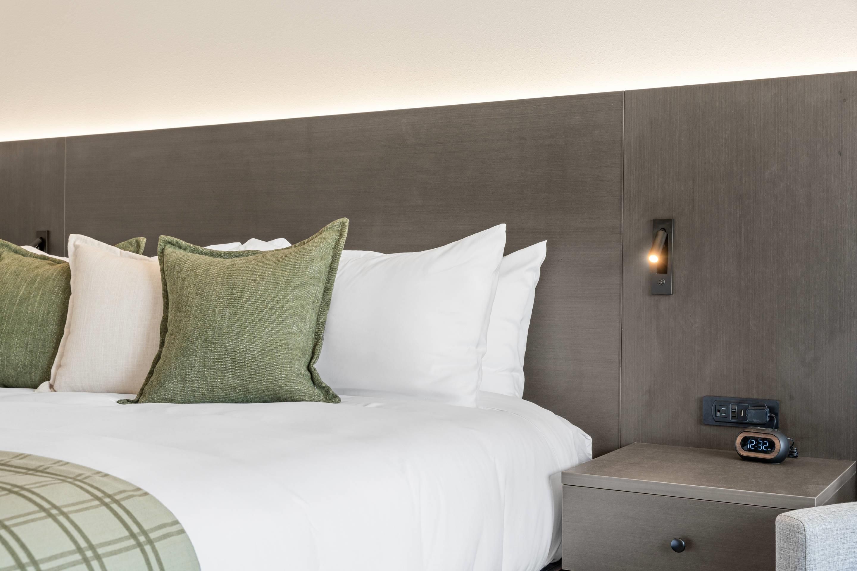 A neatly made bed with white and green pillows, a bedside table with a clock, and a wall-mounted light.