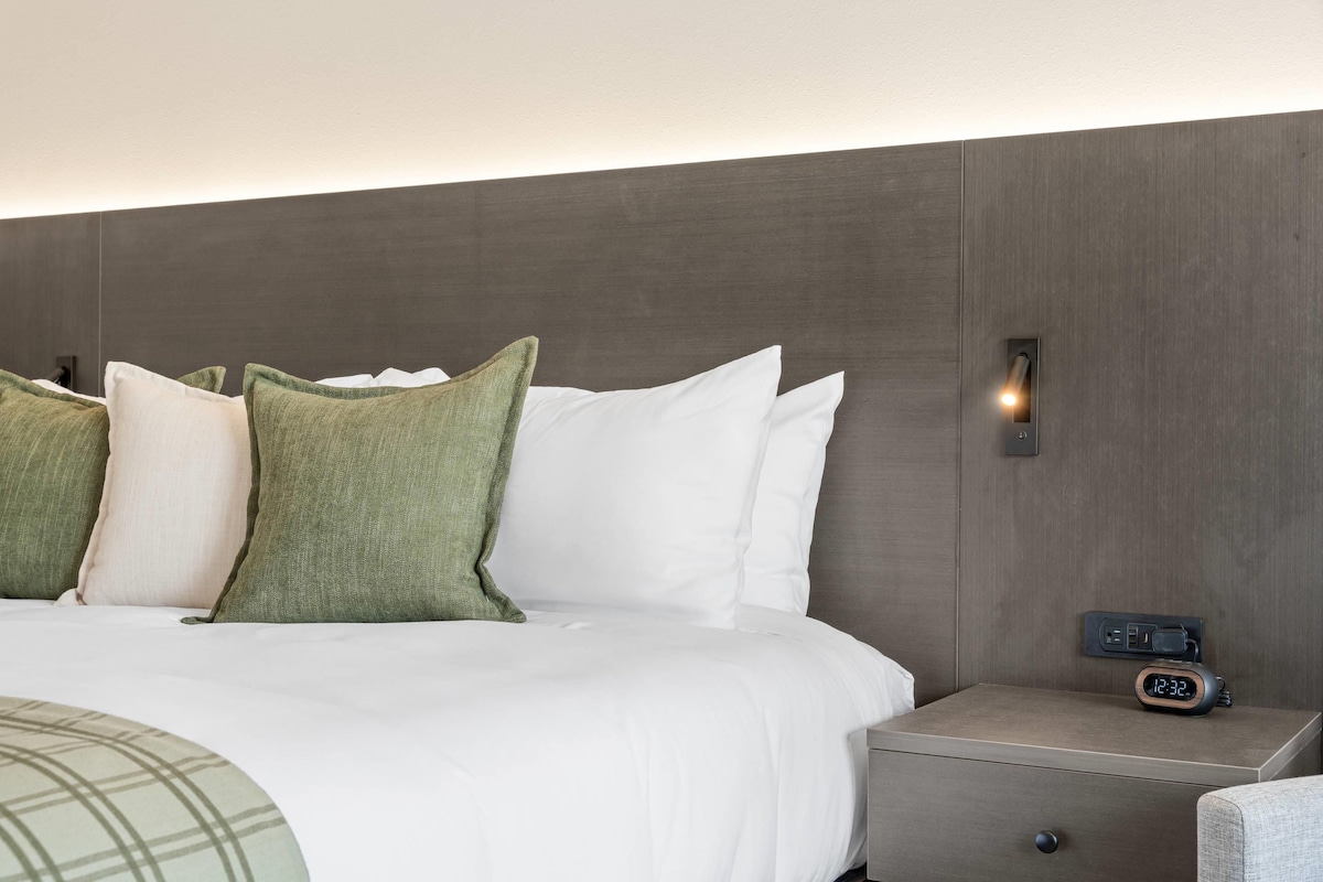 A neatly made bed with white and green pillows, a bedside table with a clock, and a wall-mounted light.