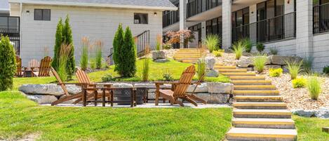 A modern building with a flat roof, outdoor seating area with wooden chairs and tables, and a landscaped garden with steps leading to the entrance.