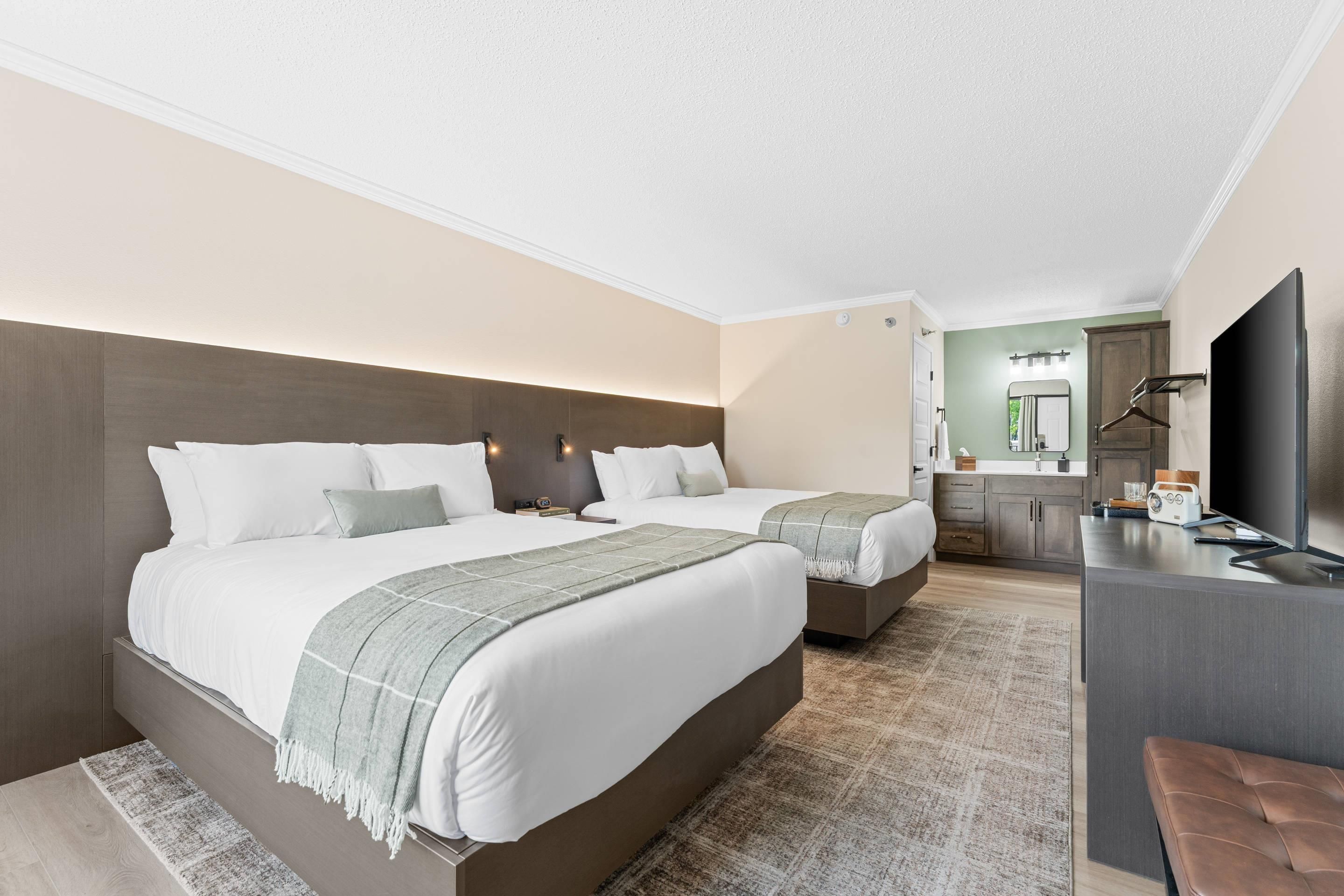 Une chambre d’hôtel avec deux lits, un bureau, une télévision et une salle de bain équipée d’un miroir et d’un lavabo.