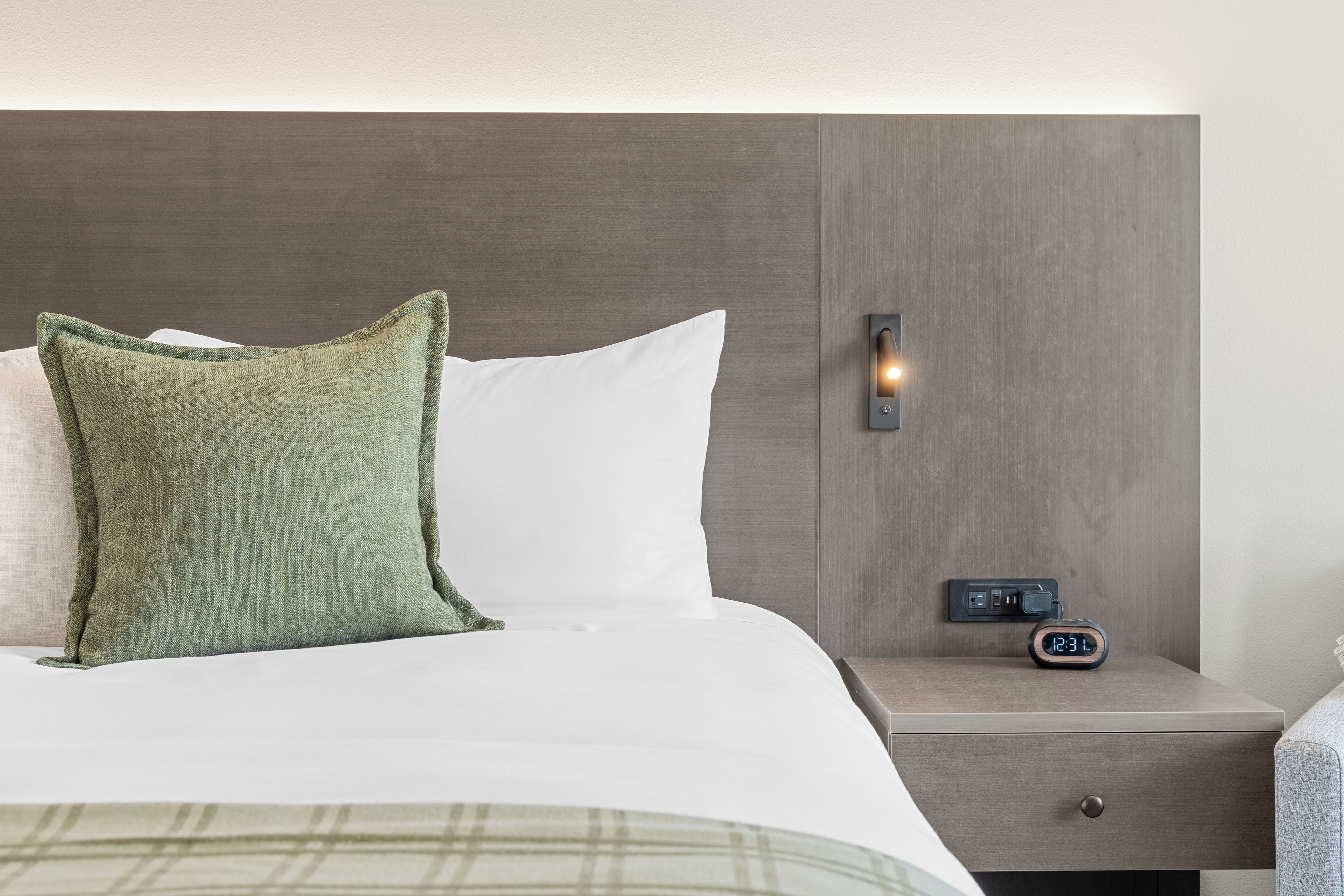 A neatly made bed with a green pillow and white pillow, a bedside table with a digital clock, and a nightstand with a drawer.