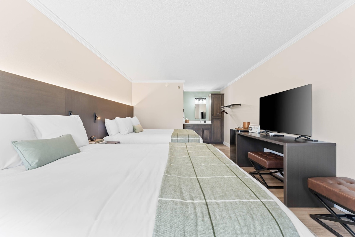 A hotel room with a bed, a desk with a television, a bench, and a bathroom visible in the background.