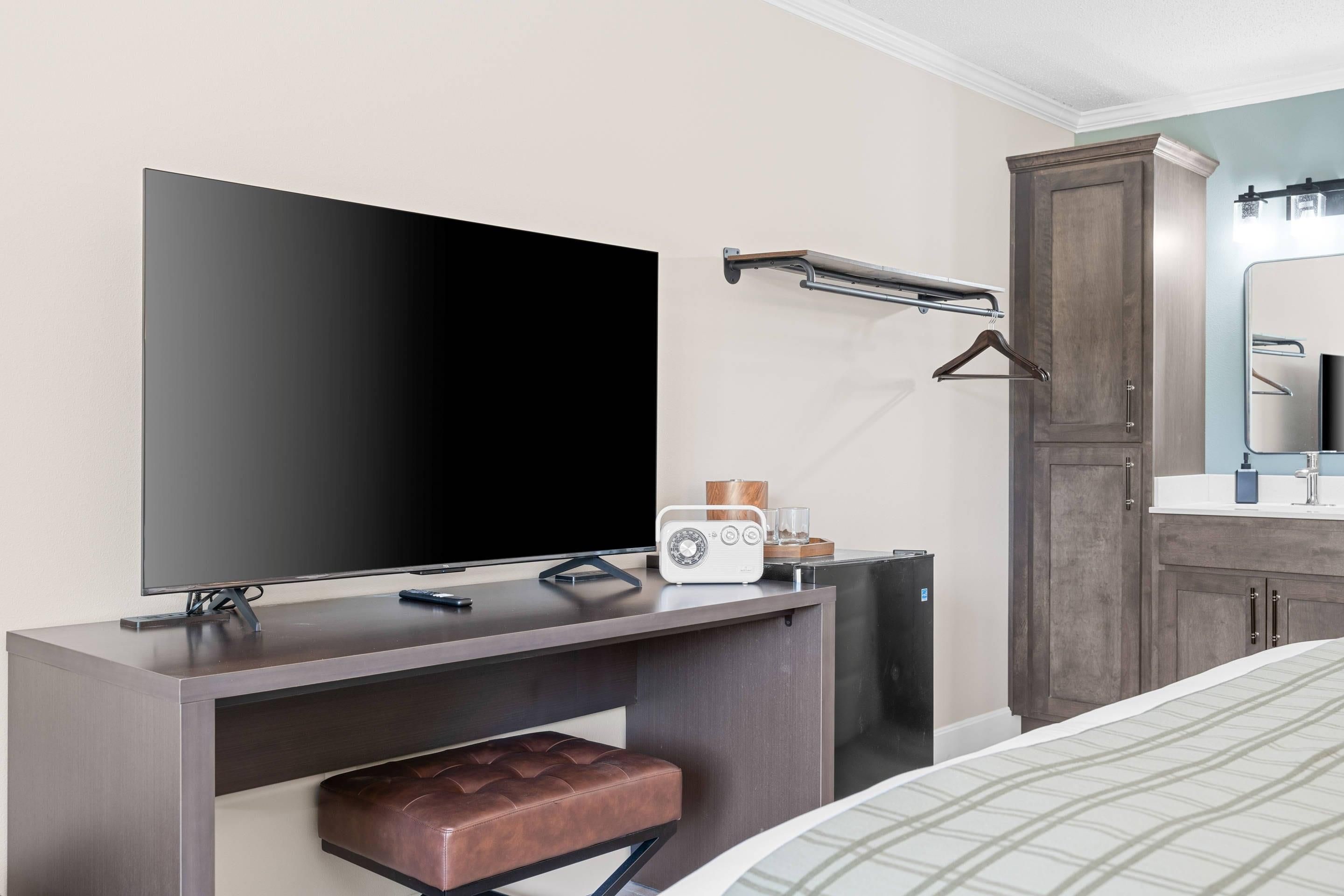 Quarto de hotel com televisão de tela plana grande, um banco de madeira, rádio, espelho e guarda-roupa.