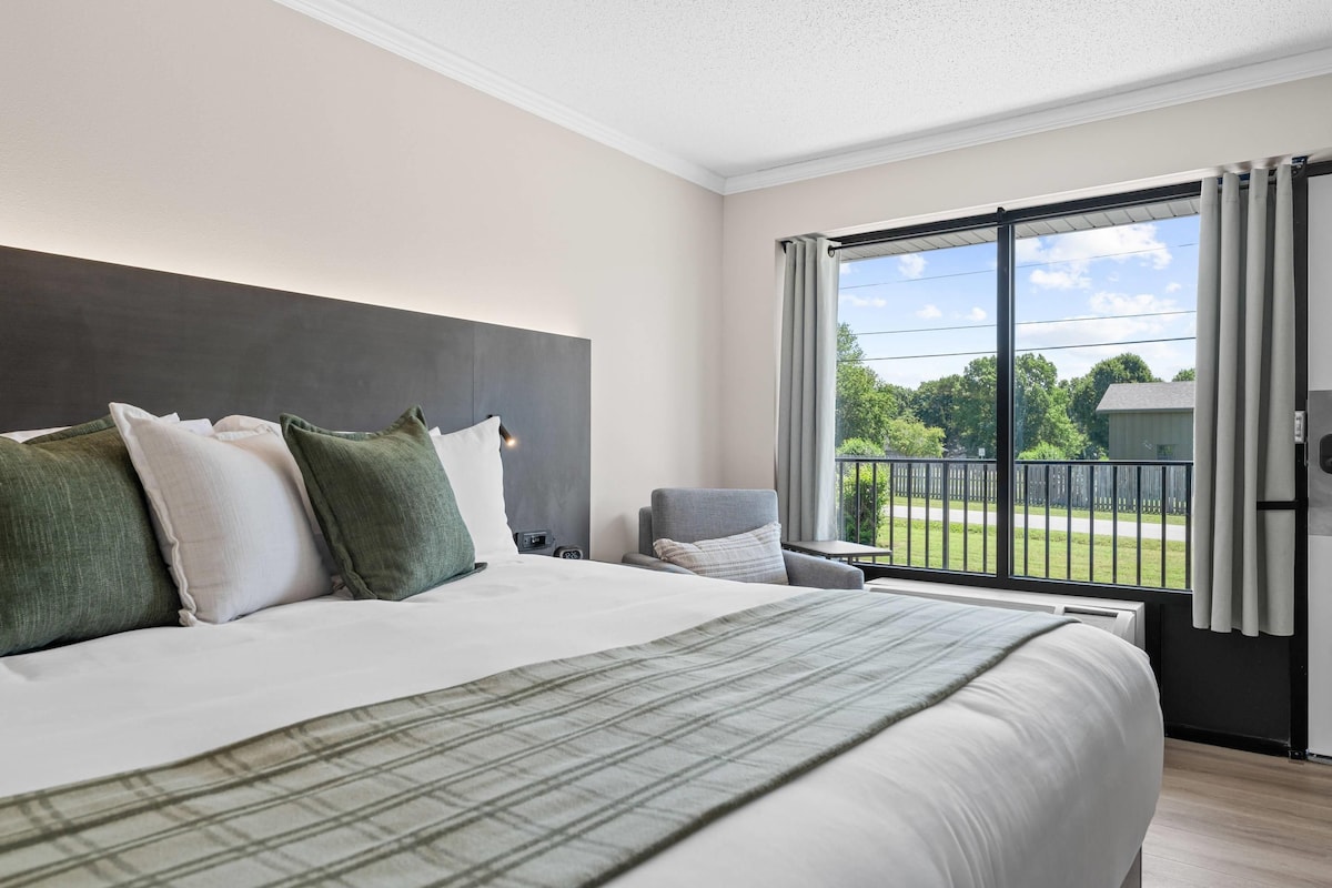 A hotel room with a large bed, a chair, a window with curtains, and a view of the outdoors.