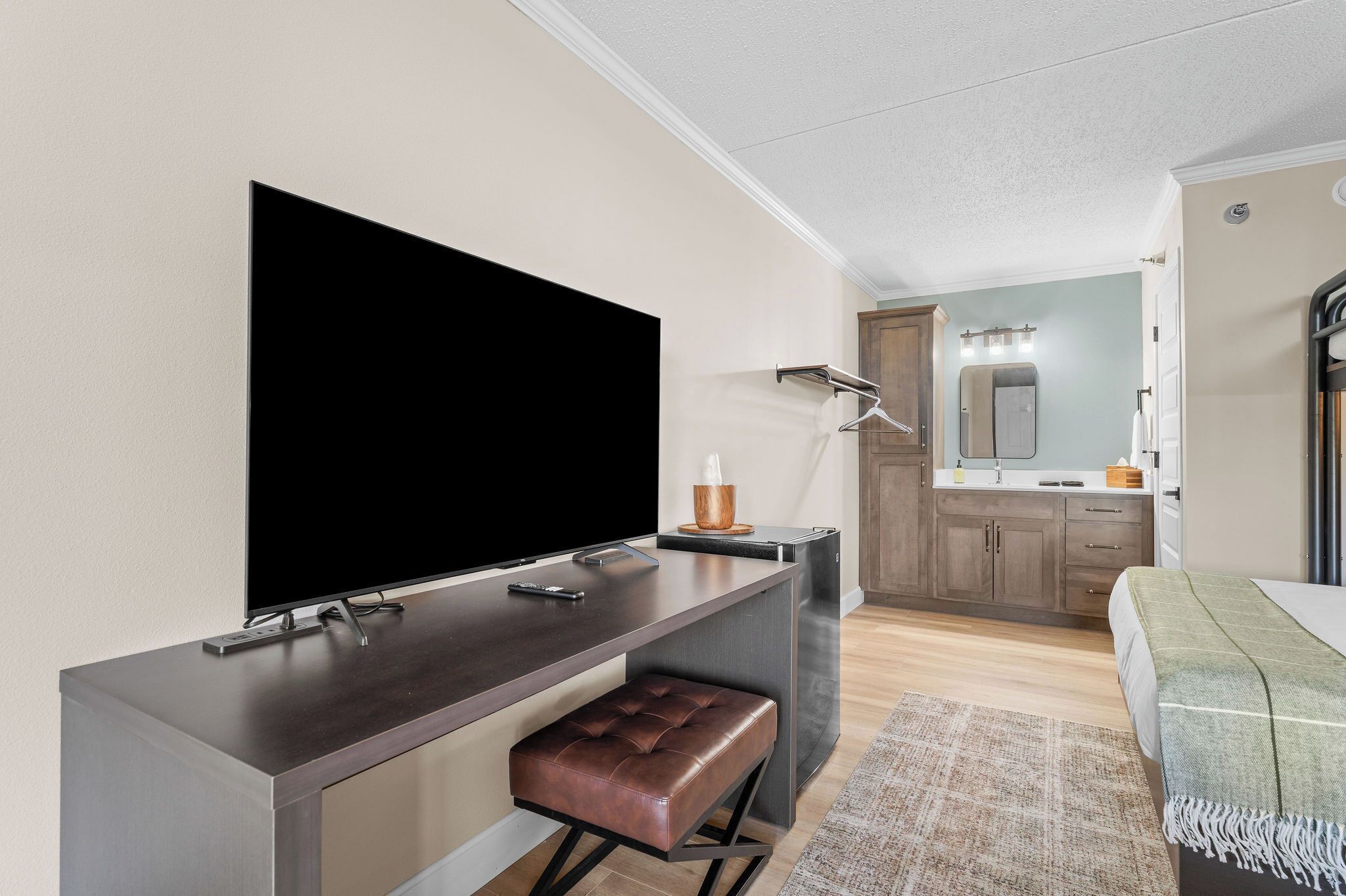 Una habitación de hotel moderna con una televisión de pantalla plana, un escritorio con una silla, un pequeño banco, un baño con espejo y una cama con manta verde.