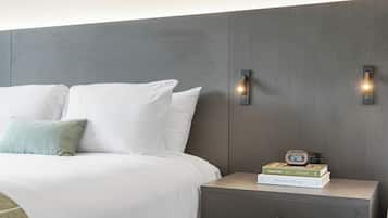 A neatly made bed with white linens and a single green pillow, a bedside table with a digital clock and books, and a wall-mounted light fixture.