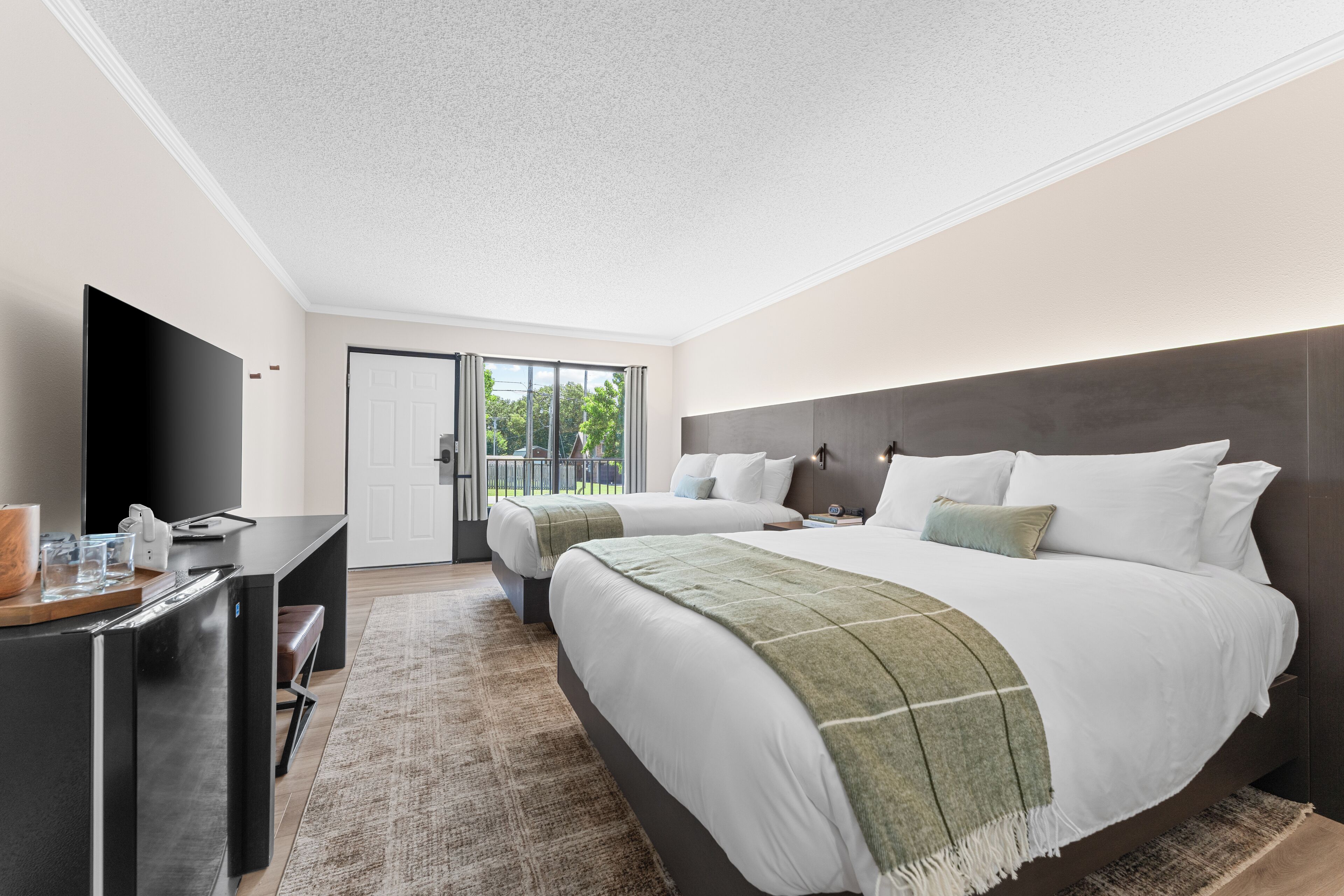 A modern hotel room with a large bed, a desk with a flat-screen TV, and a view of the outdoors through a sliding door.
