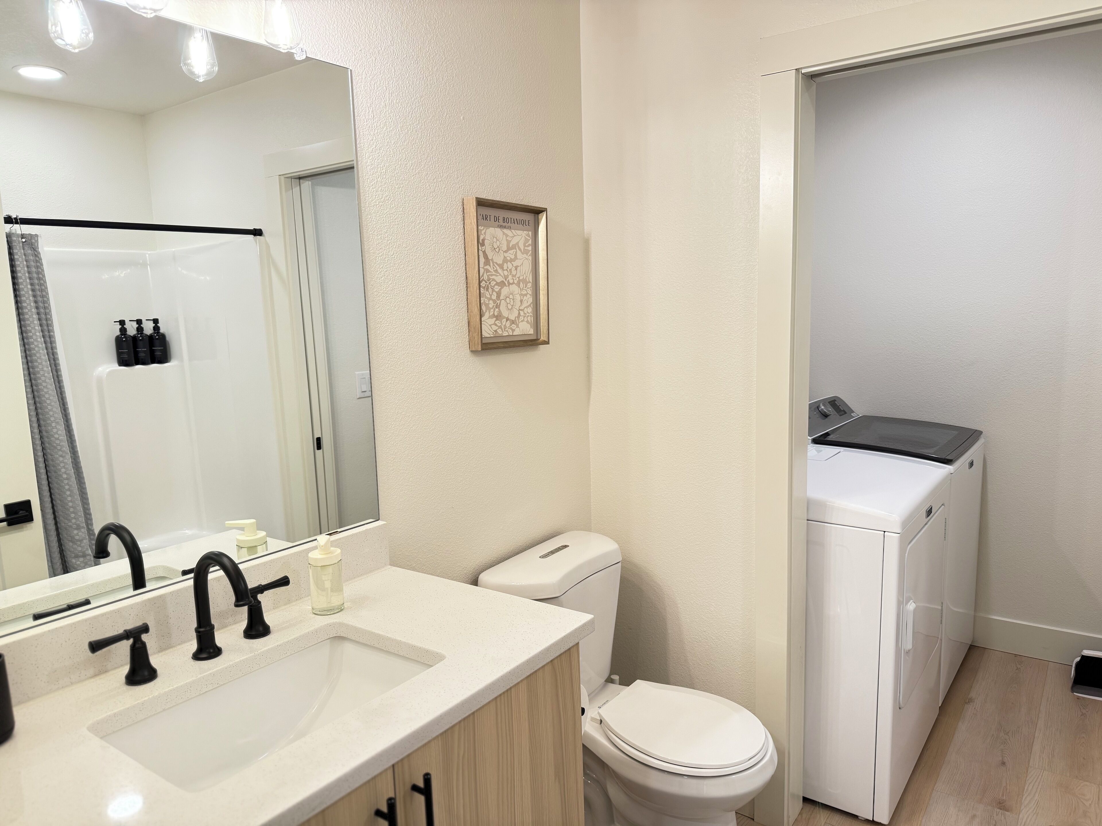 A modern bathroom with a white sink, black faucet, and a toilet. There is a shower area with a glass door and a framed picture on the wall.