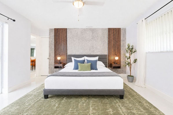 A modern bedroom with a large bed, two bedside tables with lamps, a potted plant, and a view of a dining area.