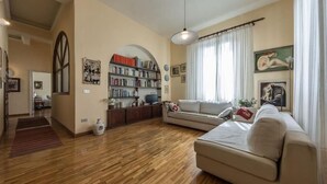 Ein geräumiges Wohnzimmer mit Holzboden, einem beigefarbenen Sofa, einem Bücherregal und einem großen Fenster mit Vorhängen.