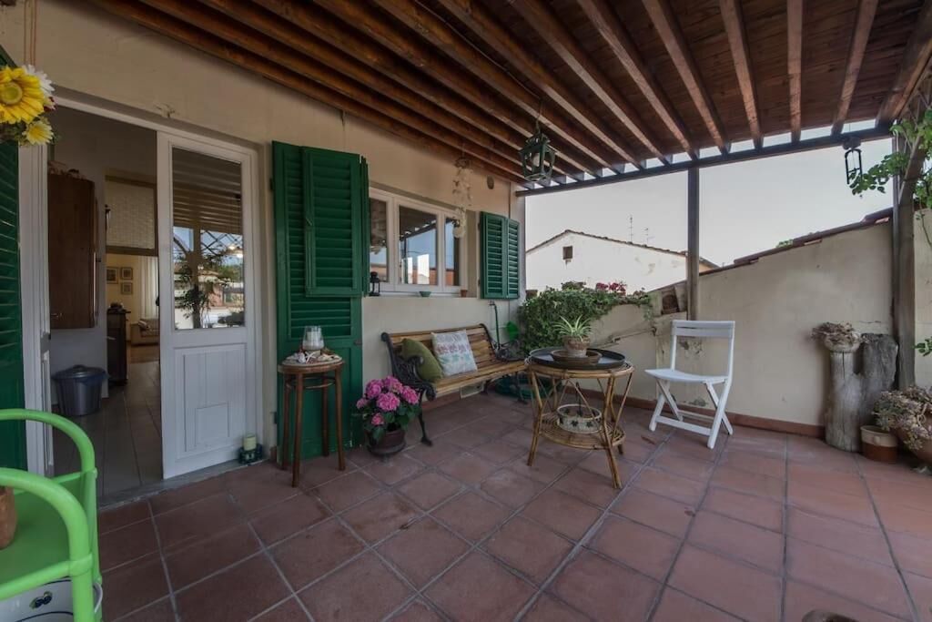 A covered patio with a wooden ceiling, green shutters, a bench, a table with a potted plant, and a white chair.