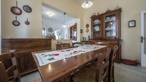 Ein Essbereich mit Holztisch, Stühlen und einer Tischdecke mit Blumenmuster. An der Wand hängen Töpfe und es gibt einen Schrank mit Glastüren.