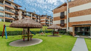 A playground with swings and a slide, surrounded by a well-maintained lawn and a building with multiple balconies. - Apt 300m From the Charming Praia Brava Ccu203 (Florianópolis)