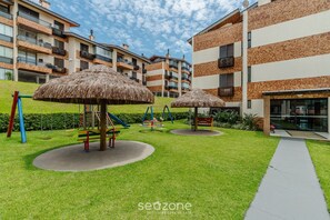 A playground with swings and a slide, surrounded by a well-maintained lawn and a building with multiple balconies. - Apt 300m From the Charming Praia Brava Ccu203 (Florianópolis)