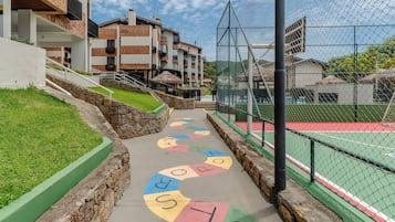 Ein farbig beletterter Weg führt zu einem Tennisplatz, umgeben von Gebäuden und einer Rasenfläche.