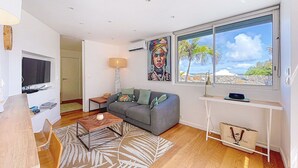 A modern living room with a grey sofa, wooden coffee table, and a large window with a view of palm trees.