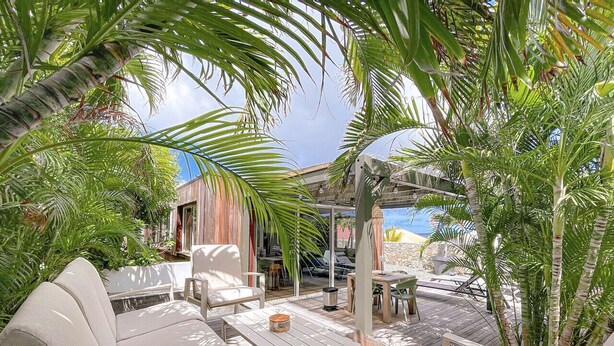 An outdoor patio area with a sofa, tables, and chairs, surrounded by lush greenery and a view of the beach.