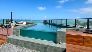 A rooftop infinity pool with a view of the ocean, lounge chairs, and a wooden deck. - Breeze (Joao Pessoa)