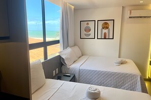 A hotel room with two beds, a window with a view of the beach, and two framed artworks on the wall.