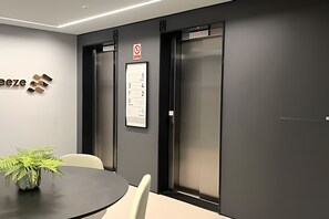 Et mødelokale med et sort bord, hvide stole, en potteplante og to elevatorer med et rygeforbudsskilte over dem.