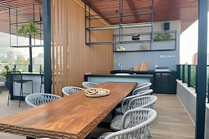 A modern outdoor dining area with a wooden table and white chairs, a kitchen area with a sink and countertop, and a view of the city skyline. - Breeze (Joao Pessoa)