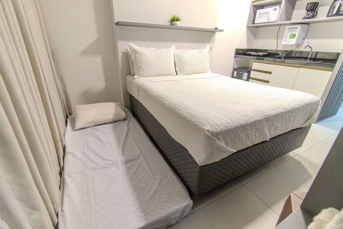 A bed with a white comforter and pillows, a bedside table, a small plant on a shelf above the bed, a microwave, and a coffee maker on the countertop.