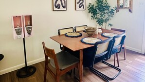 A dining area with a wooden table, four chairs, two wall-mounted abstract art pieces, a hanging light fixture, and a potted plant.