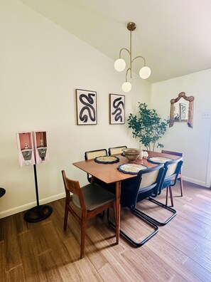A dining area with a wooden table, four chairs, two wall-mounted abstract art pieces, a hanging light fixture, and a potted plant. - Central Austin Downtown Bungalow (Austin)