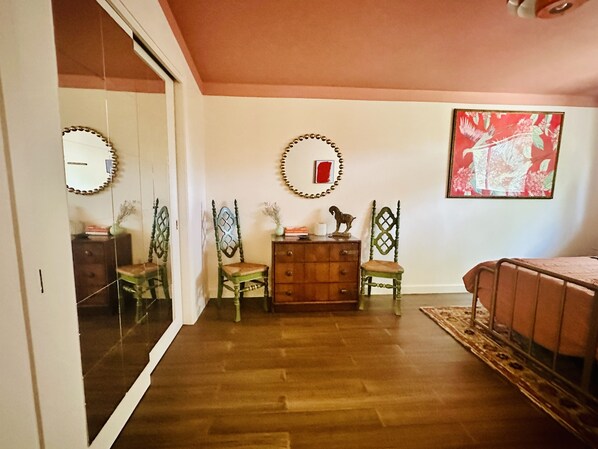 A room with wooden flooring, a mirrored closet door, two green chairs, a wooden dresser, and a red floral painting on the wall.