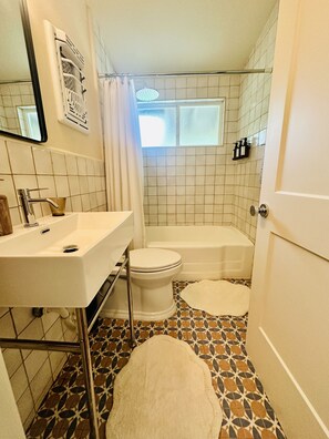A bathroom with a white sink, toilet, and bathtub. There is a window with a shower curtain, and the floor has a patterned tile design.