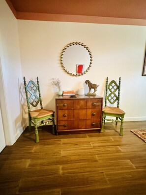 Una habitación con piso de madera, un armario de madera con cajones, dos sillas verdes, un espejo con marco dorado y un marco rojo para un cuadro en la pared.