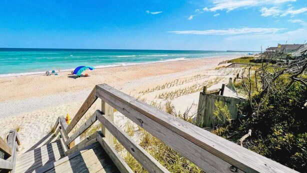 A beach with a wooden walkway leading to the shore, a beach umbrella, and a clear blue sky.