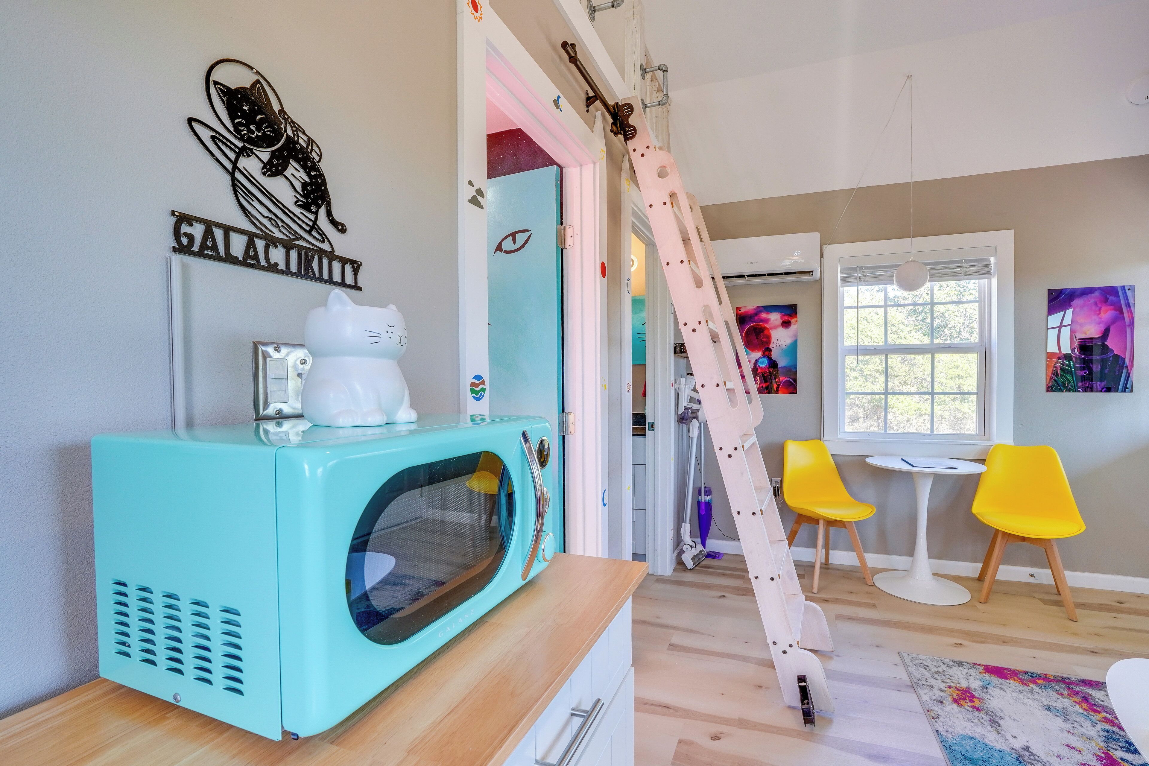 Una habitación moderna con un electrodoméstico color turquesa, una figurita de gato blanco, una escalera, sillas amarillas y un cuadro de una escena espacial.