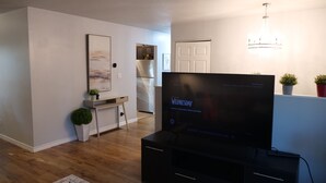 Un salon moderne avec un téléviseur à écran plat posé sur un meuble noir, un petit bureau agrémenté d’une plante, un tableau au mur et une cuisine en arrière-plan.