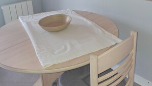 Ein runder Holztisch mit weißer Platzunterlage und einer Holzkasserolle. Ein Holzstuhl mit grauer Polsterung steht an dem Tisch. Der Raum hat hellblaue Wände und eine Holzbalkendecke.