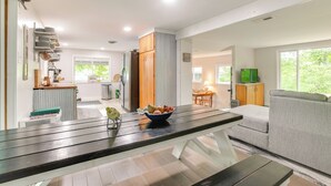 Una cocina moderna con zona de comedor, que presenta una mesa de madera con un cuenco de fruta, un sofá gris y un televisor al fondo.