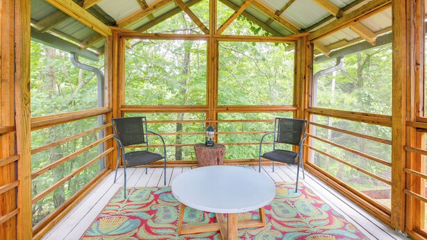 Una terraza de madera con una mesa blanca y dos sillas, rodeada de grandes ventanales que dan a un bosque.