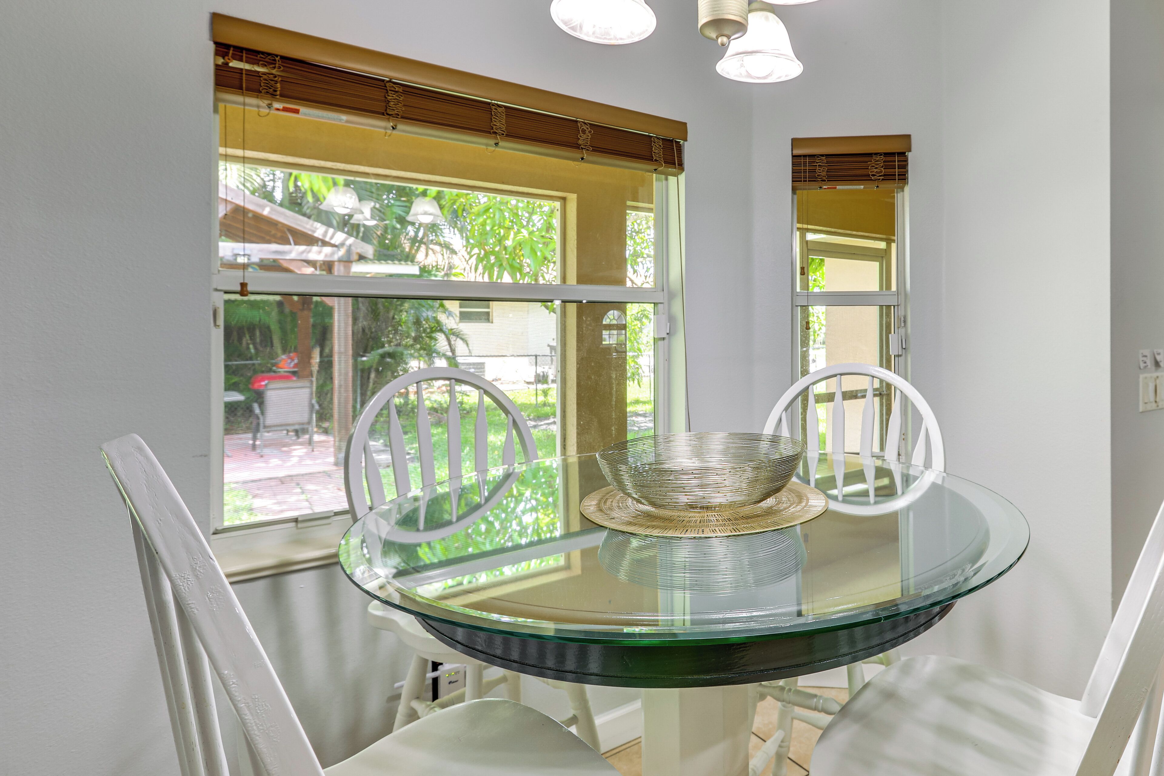 A round glass table with two white chairs, a woven basket, and a window with blinds.