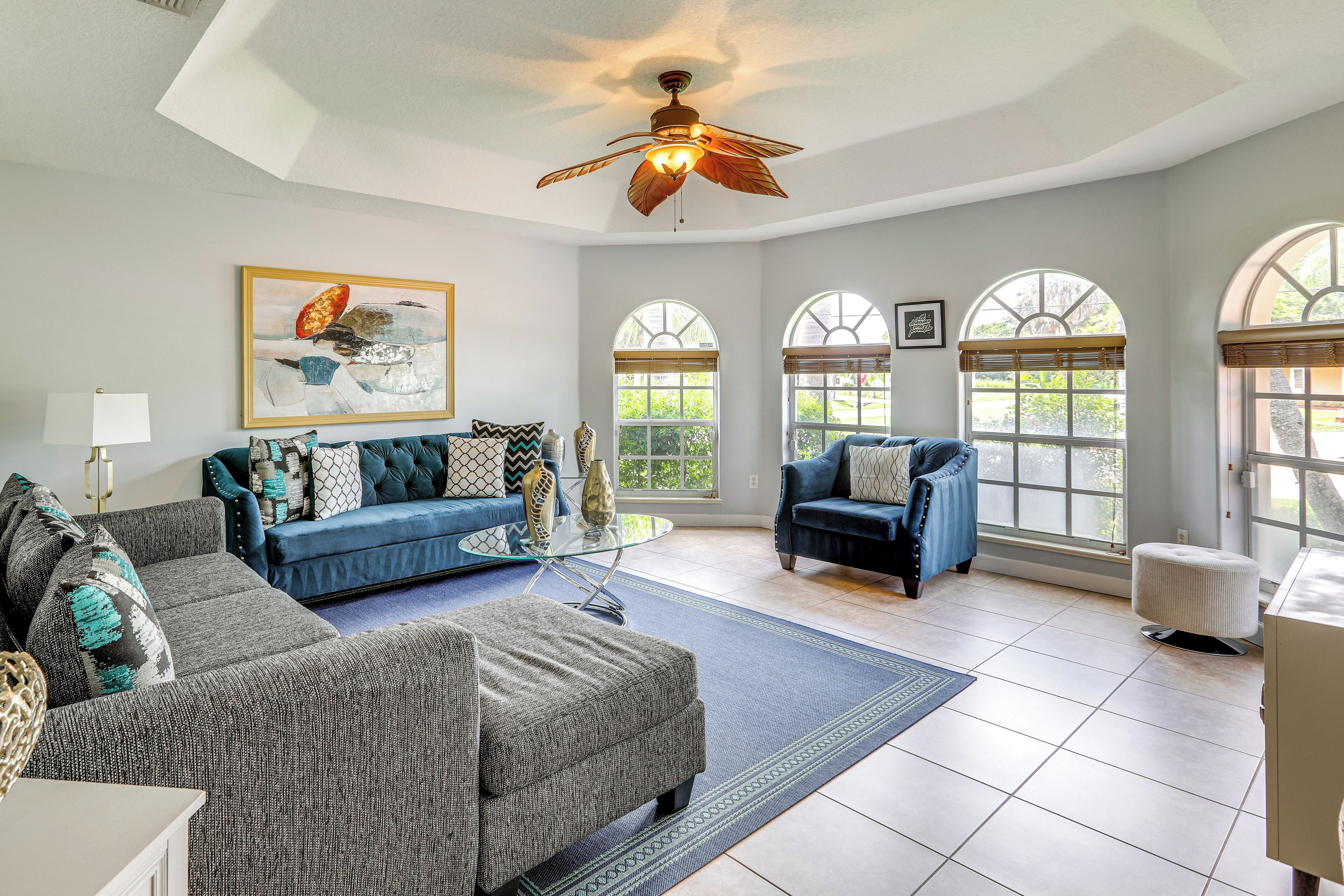 A spacious living room with a ceiling fan, a blue sofa, a patterned ottoman, and a glass coffee table.