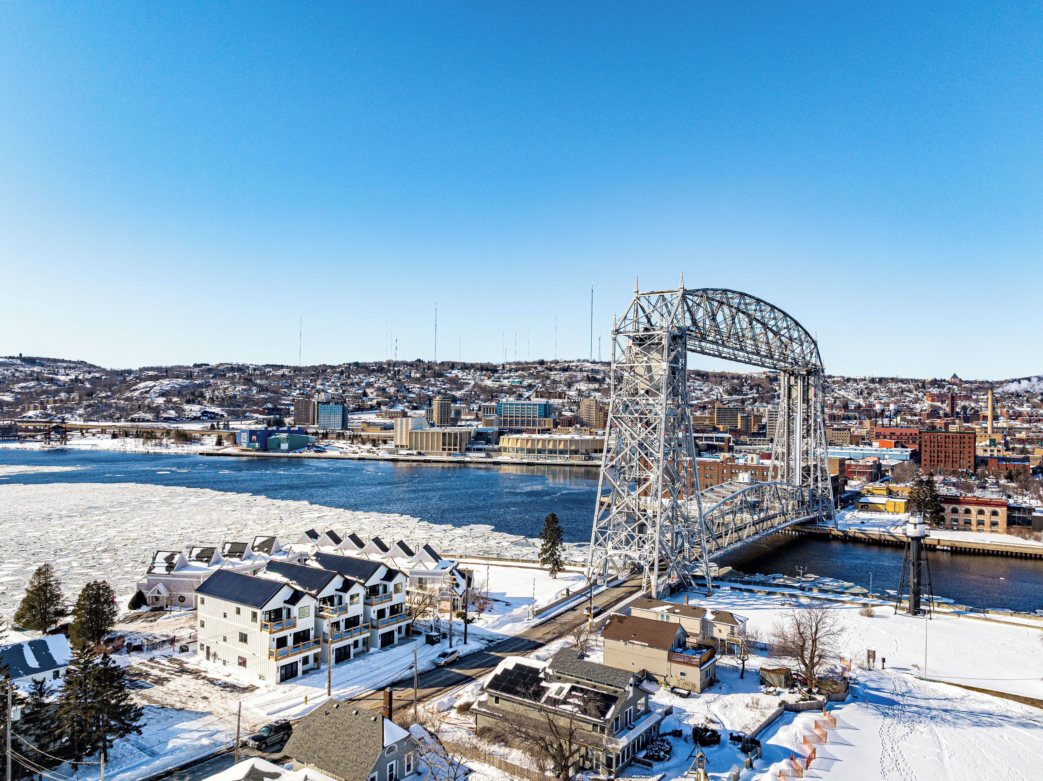 A winter scene with a large metal bridge structure, a river, and a snow-covered landscape with buildings.