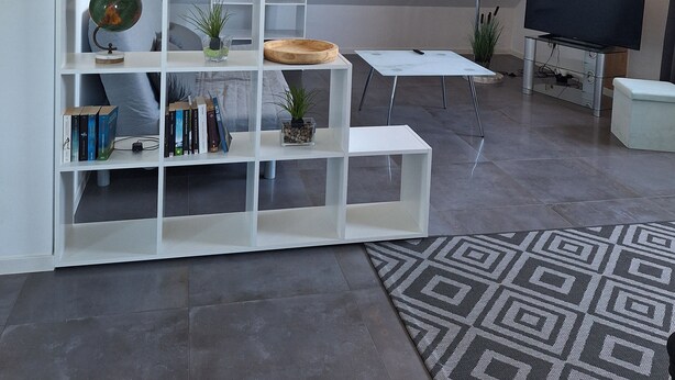 A modern living room with a sloped ceiling, white shelving units, a flat-screen TV, and a patterned rug.
